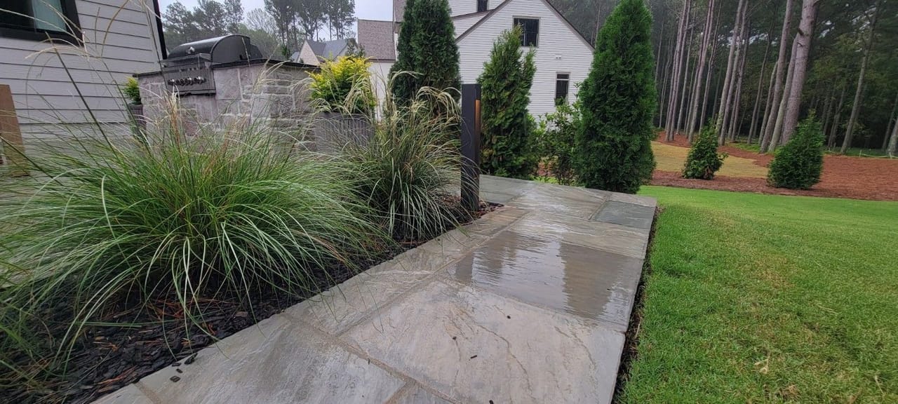Black bollard path light beside a wet flagstone walkway, grasses and outdoor kitchen on a rainy day in Wake County
