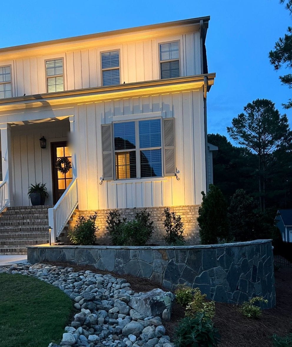 White board-and-batten farmhouse with warm up-lighting on the facade and porch at dusk in Wake County