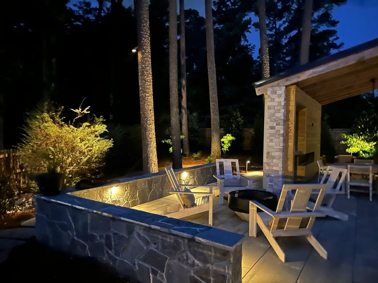 Stone seat wall with under-cap lights around a fire bowl, uplit shrubs and pines at dusk in Wake County